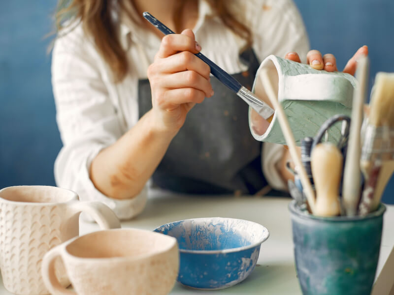 A woman surrounded by brushes and mugs paints ceramics in a class.